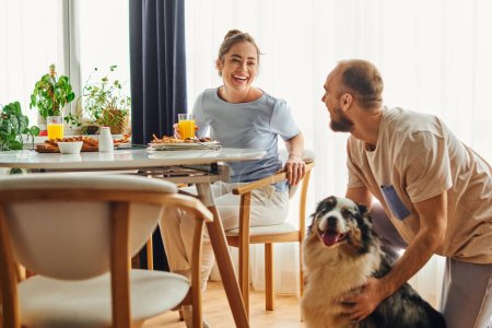 Positive woman in homewear sitting near breakfast and boyfriend with border collie at home
