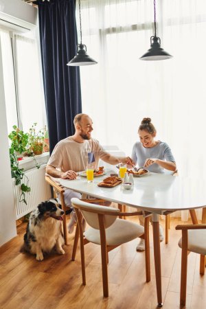 Foto de Sonriente pareja en ropa de casa desayunando juntos cerca de frontera collie perro en casa por la mañana - Imagen libre de derechos