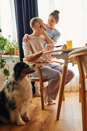 Lächelnde Frau in Homewear umarmt Freund beim Frühstück und Border Collie am Morgen zu Hause