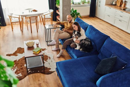 High angle view of smiling man holding remote controller near girlfriend and border collie at home