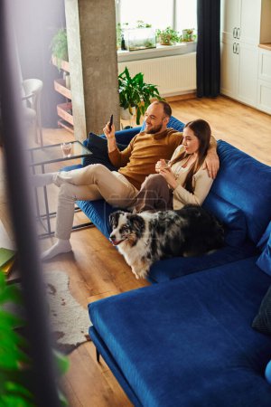Smiling man holding remote controller near girlfriend with coffee and border collie on couch at home