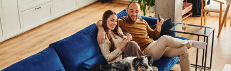 Smiling couple with coffee and remote controller sitting near border collie on couch at home,banner