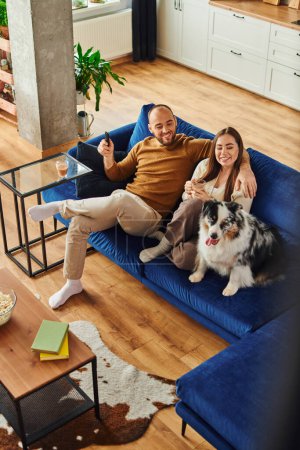 Vue grand angle du couple joyeux avec télécommande assis près de la frontière collie sur le canapé à la maison