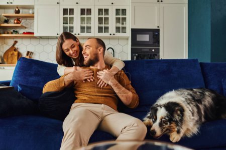 Smiling woman hugging boyfriend in casual clothes near border collie on couch in living room