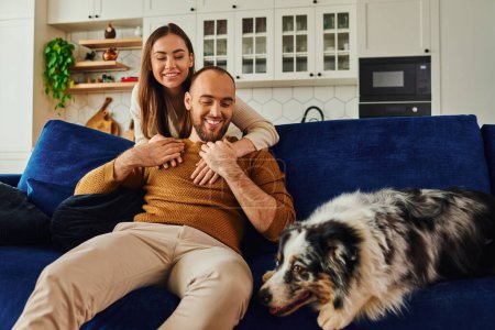 Joyful woman hugging boyfriend in casual clothes and looking at border collie on couch at home