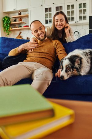 Smiling couple hugging and spending time with border collie dog on couch near books in living room