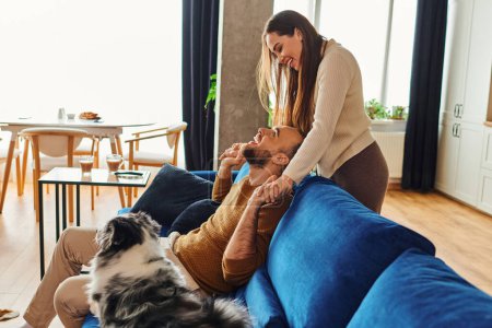 Cheerful couple in casual clothes holding hands near border collie on couch in living room at home