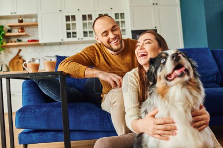 Cheerful woman hugging border collie near boyfriend and coffee in living room at home