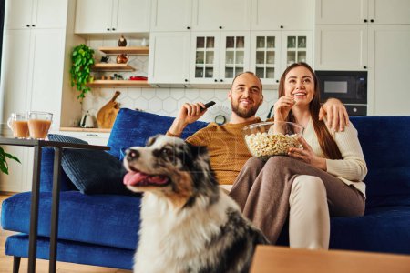 Smiling couple with remote controller and popcorn sitting near blurred border collie at home