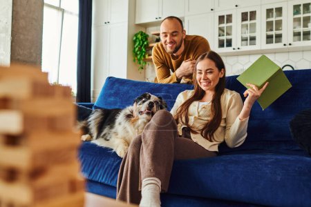 Cheerful couple with book looking at border collie on couch near blurred wood blocks game at home