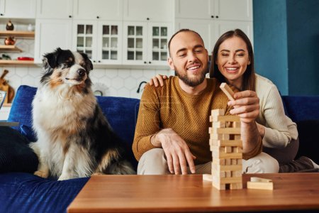 Mujer sonriente abrazando novio y jugando bloques de madera juego cerca de frontera collie perro en el sofá en casa