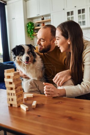 Laughing couple petting border collie while playing wood blocks game on coffee table in living room