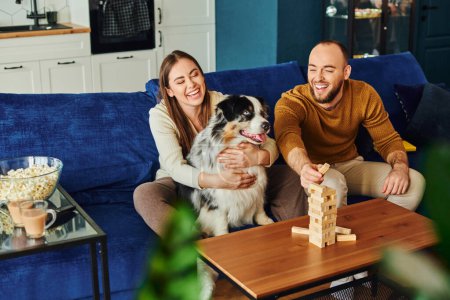 Positive couple spending time with wood blocks game and border collie on couch near popcorn at home