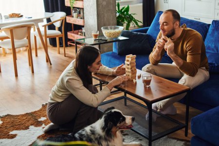 Smiling man spending time with girlfriend playing wood blocks game near border collie at home