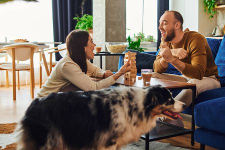 Excited couple playing wood blocks game together near border collie dog in living room at home
