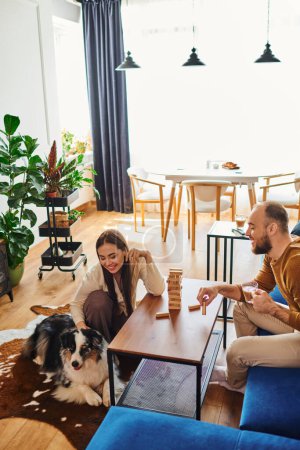 Positive couple with coffee looking at border collie while playing wood blocks game in living room