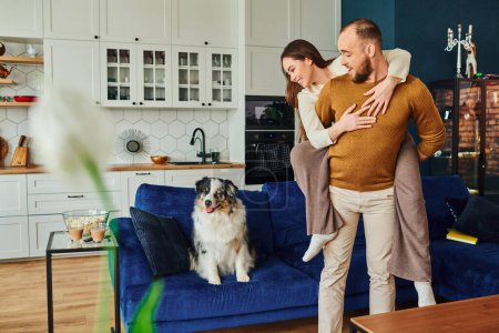 Positive woman piggybacking on boyfriend and looking at border collie on couch near popcorn at home