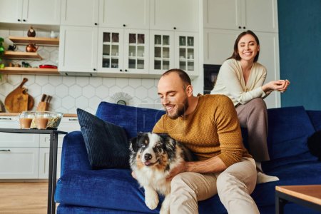 Smiling man petting border collie while sitting near girlfriend on couch and popcorn at home