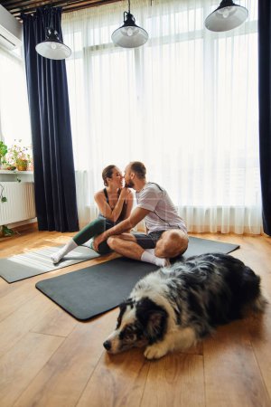 Smiling couple in sportswear kissing and sitting on fitness mats near blurred border collie at home
