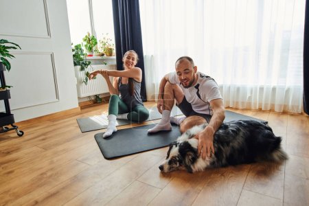 Smiling man in sportswear petting border collie while girlfriend warming up on fitness mat at home