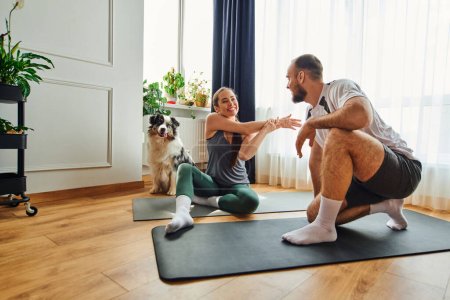 Smiling woman in sportswear warming up on fitness mat near boyfriend and border collie at home