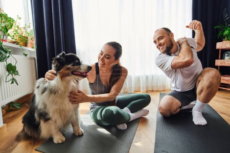 Un homme souriant s'échauffe près de sa petite amie en vêtements de sport caressant collie frontière sur tapis de fitness à la maison