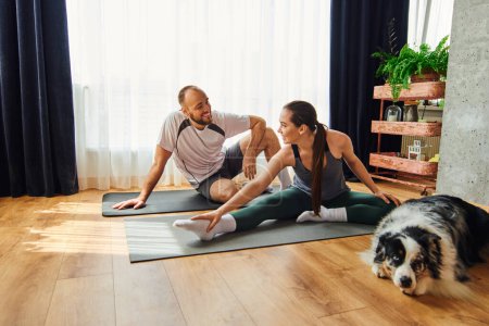 Smiling couple in sportswear talking while warming up on fitness mats near border collie at home