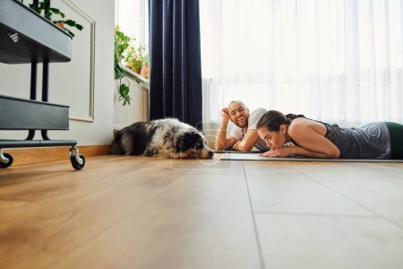 Smiling man in sportswear looking at girlfriend and border collie while lying on fitness mat at home