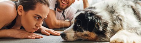 Displeased woman looking at border collie near smiling boyfriend on fitness mat at home,banner