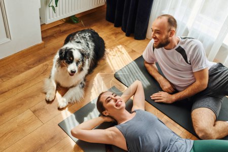 High angle view of positive couple in sportswear lying on fitness mats near border collie at home