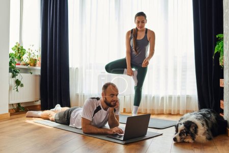 Photo for Smiling woman standing on fitness mat while boyfriend using laptop near border collie at home - Royalty Free Image