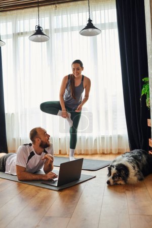 Positive woman standing on fitness mat near boyfriend using laptop and border collie at home