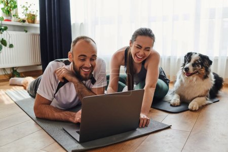 Photo for Joyful couple in sportswear using laptop together on fitness mats near border collie at home - Royalty Free Image