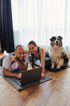 Riendo pareja en ropa deportiva usando el ordenador portátil juntos en colchonetas de fitness cerca de frontera collie perro en casa