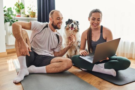 Smiling woman using laptop and sitting on fitness mat near boyfriend and border collie at home