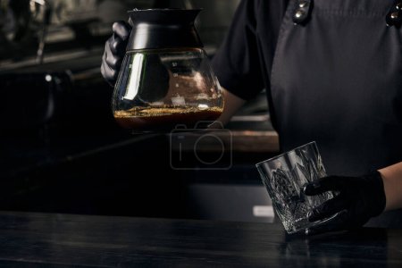 barista in black gloves holding glass and coffee pot of freshly brewed aromatic espresso
