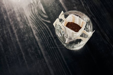 top view of aromatic ground coffee in paper filter and crystal glass on black table, pour-over brew