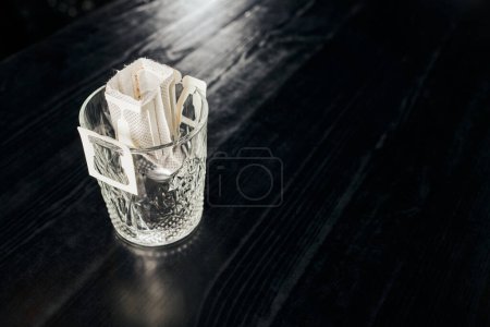 Glas mit frischem, aromatisch gemahlenem Kaffee in Filterbeutel auf schwarzer Holztischplatte, Übergießmethode