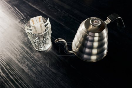 método pour-over, hervidor de agua de goteo metálico, vidrio con café molido en bolsa de filtro sobre mesa negra