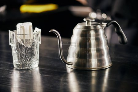 glass with ground coffee in filter bag, metallic drip kettle on black table, pour-over brewing way