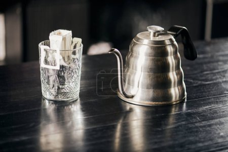 Gießverfahren, Glas mit Kaffee in Papierfilter, metallischer Tropfkessel auf schwarzem Holztisch 