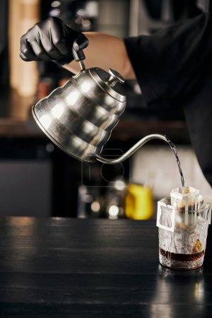 barista preparing espresso, pouring boiling water from kettle into glass with coffee in filter bag