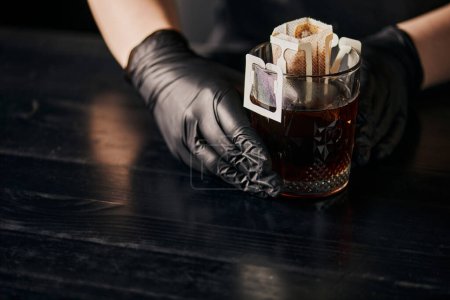 Gießverfahren, Glas mit Kaffee in Papierfilterbeutel in der Nähe von Barista in schwarzen Latex-Handschuhen 