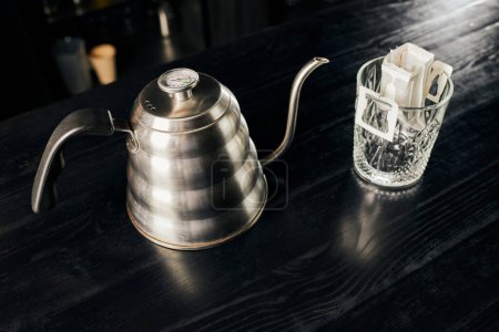 Metalowy czajnik kroplowy, szkło kryształowe z mieloną kawą w worku filtracyjnym na czarnym stole, metoda kroplowa