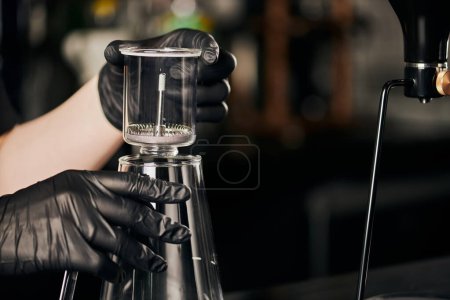 Photo for Coffee shop, barista in black latex gloves holding siphon coffee maker above glass coffee pot - Royalty Free Image