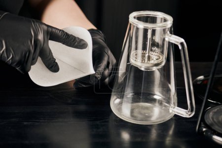 Foto de Vista recortada de barista en guantes de látex negro sosteniendo filtro de papel cerca de cafetera sifón - Imagen libre de derechos