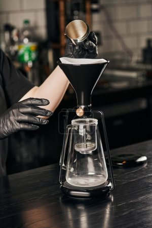 Ausgeschnittene Ansicht des Barista in schwarzen Handschuhen, der Jigger in der Nähe der zusammengebauten Siphon-Kaffeemaschine im Café hält