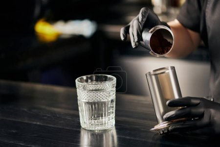 Foto de Vista parcial de barista en vidrio de látex negro sosteniendo prensa aerodinámica con café cerca de vidrio sobre mesa negra - Imagen libre de derechos