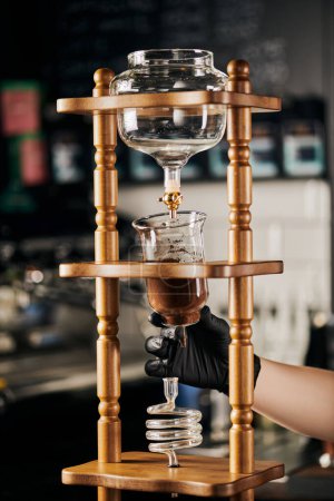 barista en guante negro ensamblando cafetera de goteo frío con café molido, cerveza espresso alternativa