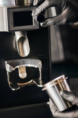 cropped view of barista in black gloves with measuring cup near electric coffee grinder, equipment